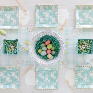 May include: A table setting with a pastel blue and white bunny patterned paper plates, a white bowl filled with pastel colored Easter eggs, and a white and pink "Happy Easter" sign.