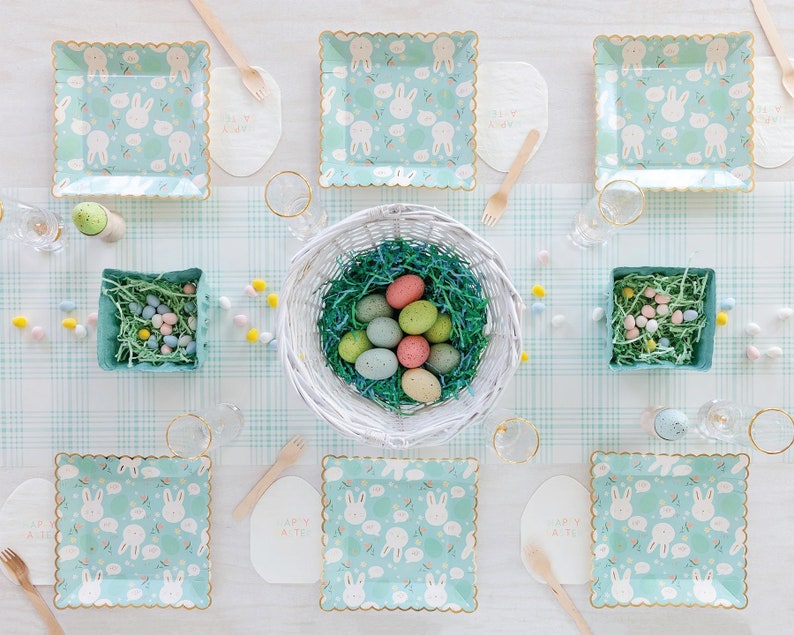 May include: A table setting with a pastel blue and white bunny patterned paper plates, a white bowl filled with pastel colored Easter eggs, and a white and pink "Happy Easter" sign.