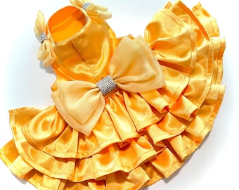 May include: A bright yellow pet dress featuring a multi-layered ruffled skirt, a large central bow with a silver rhinestone accent, and two smaller bows with rhinestone details on the neckline. The garment is made of a smooth, satin-like fabric.