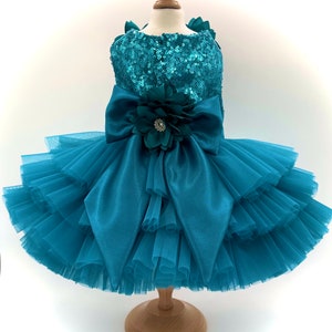May include: Teal-colored dog dress with a sequined bodice and layered tulle skirt. A large satin bow and a flower with a rhinestone center adorn the front. The dress is displayed on a wooden mannequin.
