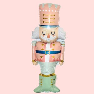 May include: A festive, pink and gold nutcracker balloon with a detailed design. The balloon features a tall hat, a smiling face, and a uniform with gold accents. The nutcracker's legs are a light green color. Perfect for holiday decorations.