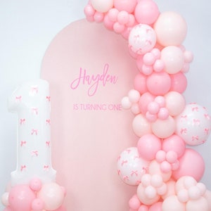 May include: A pink and white balloon arch with a large white number one balloon decorated with pink bows. The arch is in front of a pink backdrop with the text "Hayden is turning one".