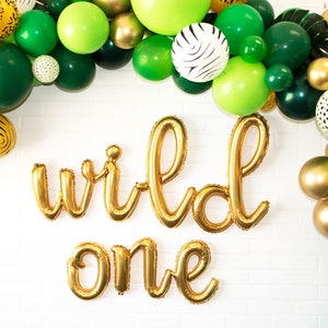 May include: Gold foil balloons spelling "wild one" with a green and gold balloon arch in the background.