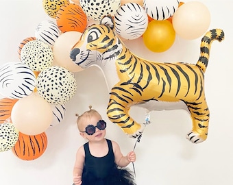 May include: A young child wearing a black tutu and sunglasses holds a large golden tiger balloon. The balloon is surrounded by a garland of colorful balloons with animal print designs.