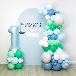 May include: A blue and green balloon arch with a large blue number one balloon in front of a light blue backdrop. The backdrop has the text 