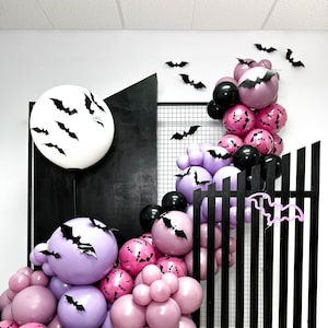 May include: A Halloween-themed balloon arch featuring a large white balloon with black bat cutouts, and a cascade of purple, pink, and black balloons. The balloons are decorated with bat silhouettes. A black wooden structure and bat cutouts complete the festive display.
