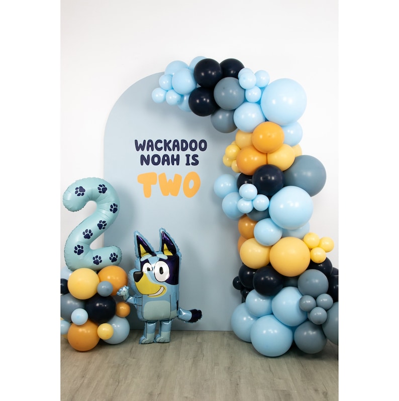 Puede incluir: Un arco de globos azul y amarillo con el texto "Wackadoo Noah is Two" sobre un fondo azul. Un globo de perro azul y un globo grande con el n&uacute;mero dos est&aacute;n en primer plano.