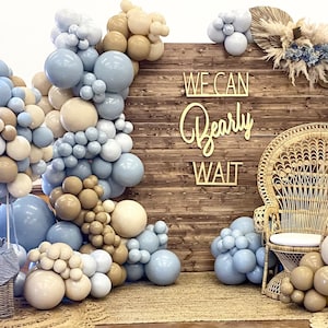 Puede incluir: Un tel&oacute;n de fondo de madera r&uacute;stica con un letrero que dice "We Can Bearly Wait". El tel&oacute;n de fondo est&aacute; decorado con globos azules y beige, una silla de mimbre y un oso de peluche en una cesta.