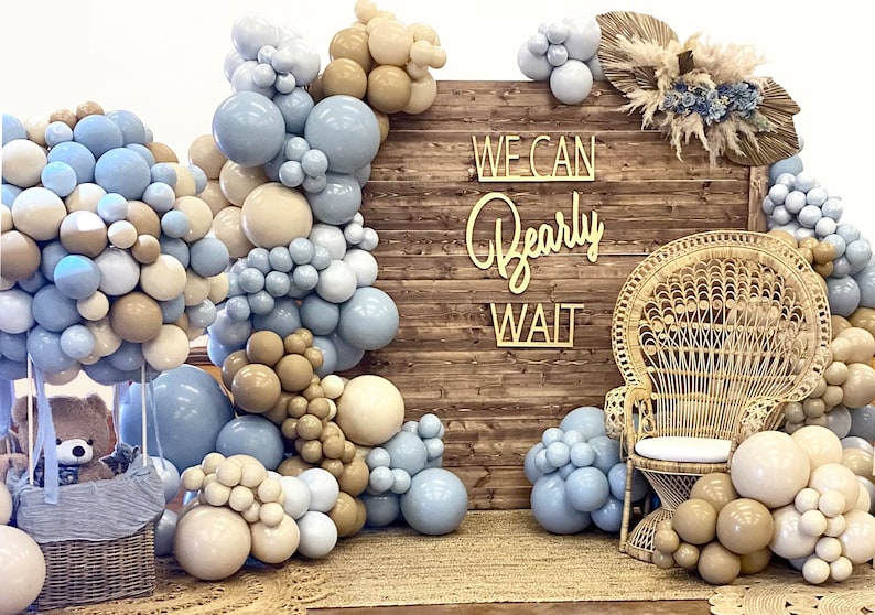 Puede incluir: Un tel&oacute;n de fondo de madera r&uacute;stica con un letrero que dice "We Can Bearly Wait". El tel&oacute;n de fondo est&aacute; decorado con globos azules y beige, una silla de mimbre y un oso de peluche en una cesta.