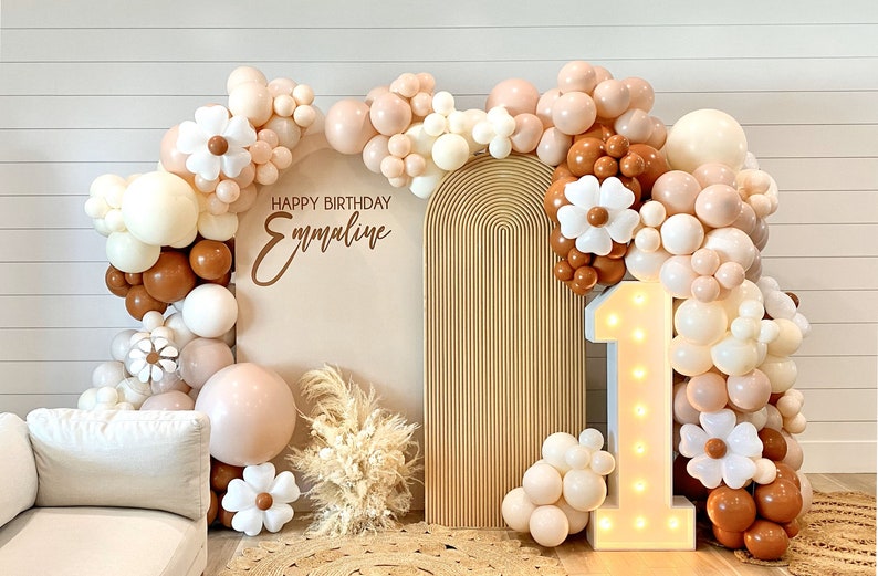 Puede incluir: Un tel&oacute;n de fondo para una fiesta de primer cumplea&ntilde;os con un arco de color marr&oacute;n claro, un n&uacute;mero uno blanco iluminado y una guirnalda de globos en tonos marrones, blancos y crema. La guirnalda de globos est&aacute; decorada con flores de papel blancas. El tel&oacute;n de fondo tambi&eacute;n incluye un cartel que dice "Feliz cumplea&ntilde;os Emmaline".