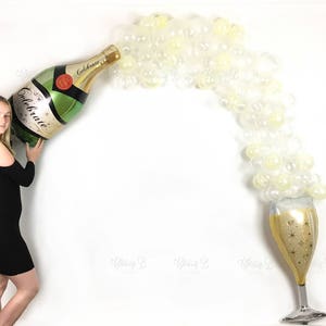 May include: A large gold and green inflatable champagne bottle with the word "Celebrate" on the label. The bottle is being held by a person. A stream of clear balloons flows from the bottle's spout into a gold champagne glass.