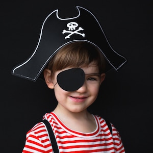 May include: A child wearing a black pirate hat with a white skull and crossbones, a black eye patch, and a red and white striped shirt. The background is solid black.