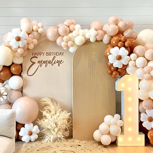 Puede incluir: Un tel&oacute;n de fondo para una fiesta de primer cumplea&ntilde;os con un arco de color marr&oacute;n claro, un n&uacute;mero uno blanco iluminado y una guirnalda de globos en tonos marrones, blancos y crema. La guirnalda de globos est&aacute; decorada con flores de papel blancas. El tel&oacute;n de fondo tambi&eacute;n incluye un cartel que dice "Feliz cumplea&ntilde;os Emmaline".