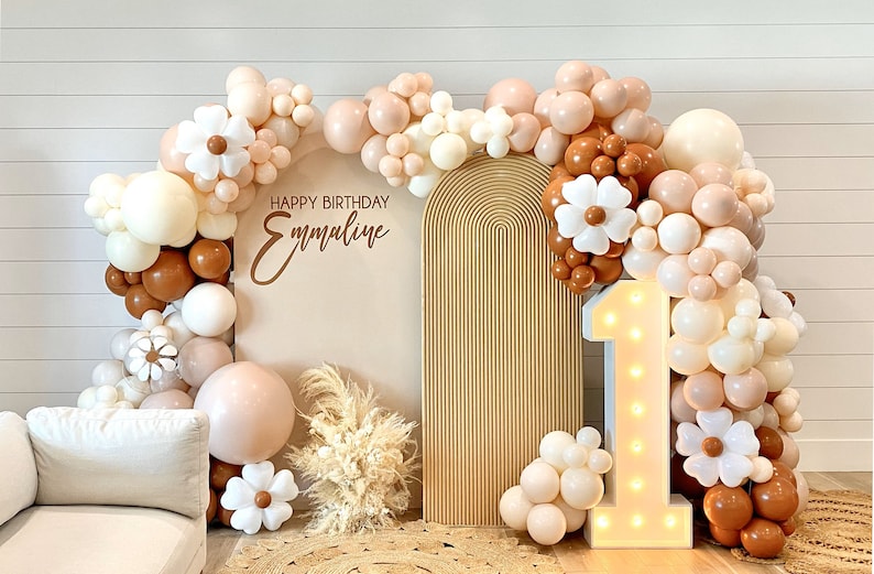 Puede incluir: Un tel&oacute;n de fondo para una fiesta de primer cumplea&ntilde;os con un arco de color marr&oacute;n claro, un n&uacute;mero uno blanco iluminado y una guirnalda de globos en tonos marrones, blancos y crema. La guirnalda de globos est&aacute; decorada con flores de papel blancas. El tel&oacute;n de fondo tambi&eacute;n incluye un cartel que dice "Feliz cumplea&ntilde;os Emmaline".