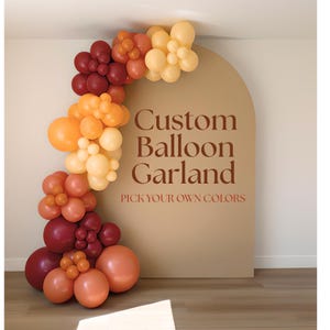 May include: A balloon garland featuring a mix of burgundy, orange, peach, and cream balloons arranged in clusters along the side of a tan arched backdrop. The backdrop displays the text Custom Balloon Garland and PICK YOUR OWN COLORS.