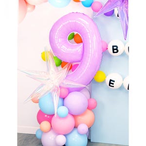 May include: A purple number nine balloon with a star-shaped balloon and a cluster of pastel-colored balloons. The balloons are arranged in a tower shape. The background is a light blue wall with a white banner that says "BE" in black letters.