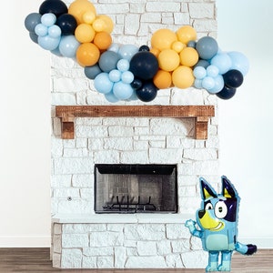 Puede incluir: Una guirnalda de globos azules y amarillos cuelga sobre una chimenea de ladrillo blanco. Un globo de perro de dibujos animados azul se encuentra frente a la chimenea.
