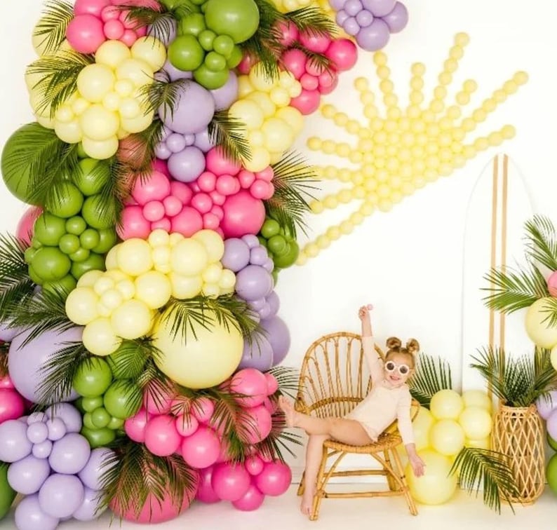 May include: A child sits in a wicker chair in front of a balloon archway. The archway is made of green, yellow, pink, and purple balloons. There is a yellow balloon sun behind the child.