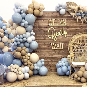 Puede incluir: Un tel&oacute;n de fondo de madera r&uacute;stica con un letrero que dice "We Can Bearly Wait". El tel&oacute;n de fondo est&aacute; decorado con globos azules y beige, una silla de mimbre y un oso de peluche en una cesta.