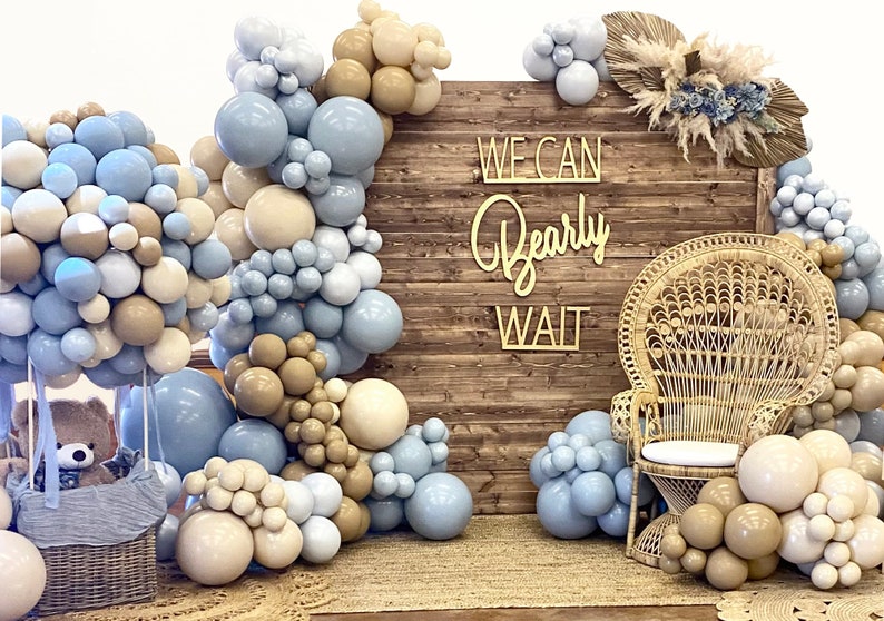 Puede incluir: Un tel&oacute;n de fondo de madera r&uacute;stica con un letrero que dice "We Can Bearly Wait". El tel&oacute;n de fondo est&aacute; decorado con globos azules y beige, una silla de mimbre y un oso de peluche en una cesta.