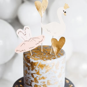 May include: A two-tiered cake decorated with gold leaf and topped with paper cutouts of a swan, a heart, and a ballerina. The cake is on a gray background.