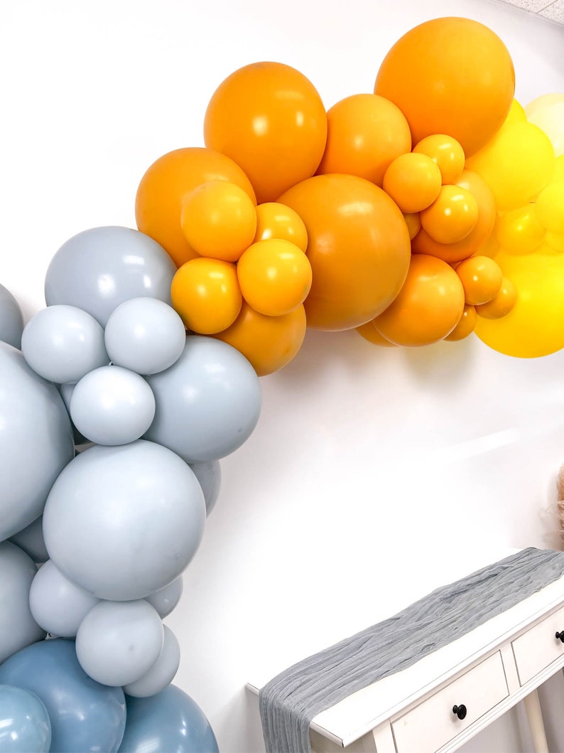 May include: A balloon arch made of light blue and yellow balloons. The arch is decorated with a gray table runner.