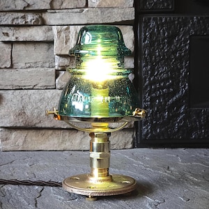 Puede incluir: Una lámpara de mesa única hecha de un aislador de vidrio verde vintage. La lámpara presenta herrajes de latón y una base redonda. El texto "MADE IN USA" es visible. La lámpara está encendida, emitiendo un brillo cálido.