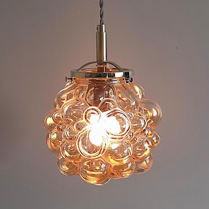 Pode incluir: Uma luminária pendente com um conjunto de abajures de vidro em forma de bolha na cor âmbar quente. A luminária tem uma estrutura de metal cor de latão e um cabo cinza. A luz está acesa, iluminando as esferas de vidro.