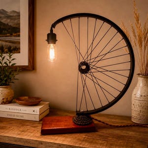 Può includere: Una lampada da tavolo costruita con un cerchio e raggi di bicicletta neri, montata su una base rettangolare in legno. La lampada presenta una singola lampadina in stile Edison appesa alla parte superiore del cerchio, collegata da un cavo intrecciato marrone.