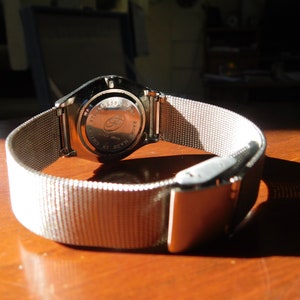 Op de afbeelding: Close-up van een zilveren polshorloge met een mesh band. De achterkant van de horlogekast is zichtbaar, met details. De band is vastgezet met een zilveren sluiting. Het horloge ligt op een houten oppervlak.