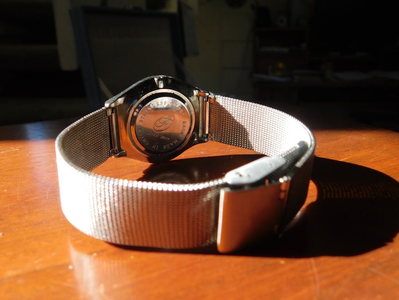 Op de afbeelding: Close-up van een zilveren polshorloge met een mesh band. De achterkant van de horlogekast is zichtbaar, met details. De band is vastgezet met een zilveren sluiting. Het horloge ligt op een houten oppervlak.