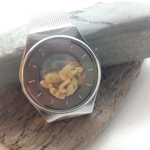 Op de afbeelding: Een zilverkleurig horloge met een mesh band en een donkergrijze wijzerplaat. De wijzerplaat bevat een verzameling kleine, lichtgekleurde, organisch gevormde objecten. Het horloge ligt op een stuk drijfhout en een grijze steen.