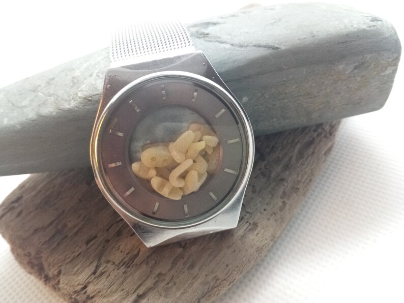 Op de afbeelding: Een zilverkleurig horloge met een mesh band en een donkergrijze wijzerplaat. De wijzerplaat bevat een verzameling kleine, lichtgekleurde, organisch gevormde objecten. Het horloge ligt op een stuk drijfhout en een grijze steen.