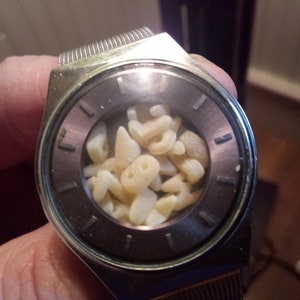 Op de afbeelding: Een zilverkleurig polshorloge met een donkergrijze wijzerplaat en een helder kristal. De wijzerplaat van het horloge is gevuld met kleine, lichtgekleurde, onregelmatig gevormde objecten. Het horloge heeft een metalen band.