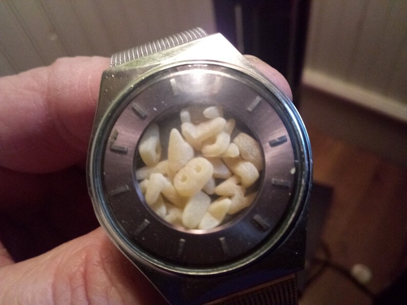Op de afbeelding: Een zilverkleurig polshorloge met een donkergrijze wijzerplaat en een helder kristal. De wijzerplaat van het horloge is gevuld met kleine, lichtgekleurde, onregelmatig gevormde objecten. Het horloge heeft een metalen band.