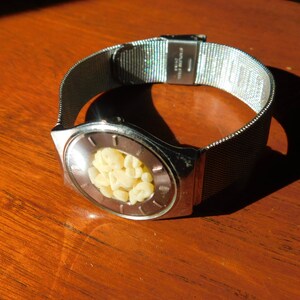 Op de afbeelding: Een zilverkleurig polshorloge met een mesh band en een unieke wijzerplaat. De ovale wijzerplaat bevat een cluster van kleine, cr&egrave;mekleurige objecten. Het horloge ligt op een houten oppervlak met schaduwen.