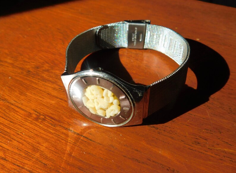Op de afbeelding: Een zilverkleurig polshorloge met een mesh band en een unieke wijzerplaat. De ovale wijzerplaat bevat een cluster van kleine, cr&egrave;mekleurige objecten. Het horloge ligt op een houten oppervlak met schaduwen.