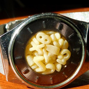 Op de afbeelding: Een zilveren polshorloge met een donkere wijzerplaat en een mesh band. De wijzerplaat bevat een verzameling gele, lettervormige pasta. Het horloge ligt op een houten oppervlak.