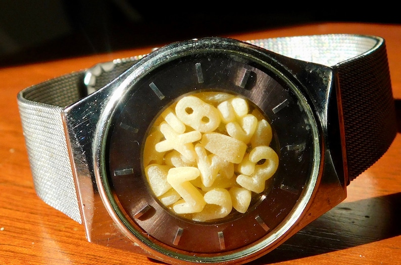 Op de afbeelding: Een zilveren polshorloge met een donkere wijzerplaat en een mesh band. De wijzerplaat bevat een verzameling gele, lettervormige pasta. Het horloge ligt op een houten oppervlak.