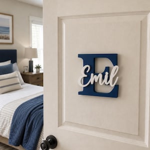 May include: A navy blue wooden letter E mounted on a white door, featuring the name Emil in white cursive script lettering layered over the centre of the letter. The sign is positioned above a black round door knob on a panelled door surface.