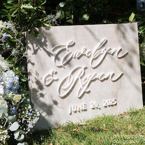 May include: A rectangular sign bearing the names "Evelyn & Ryan" in white script, with "June 20, 2025" below. The sign is placed on grass, surrounded by foliage and flowers, indicating a wedding or special occasion.
