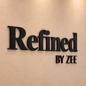 May include: A three-dimensional wall sign featuring the black text Refined BY ZEE mounted on a smooth, light beige wall. The lettering has a clean, bold typeface with a slight shadow effect, creating a professional and clear display for the brand name.