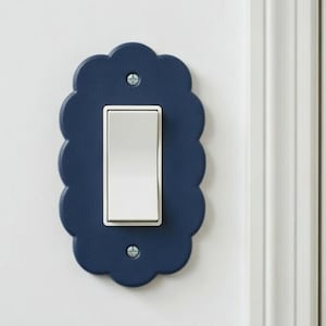 May include: A navy blue light switch cover with a scalloped edge. The cover has a rectangular opening for the white light switch and is secured to the wall with two screws. The switch is in the on position.
