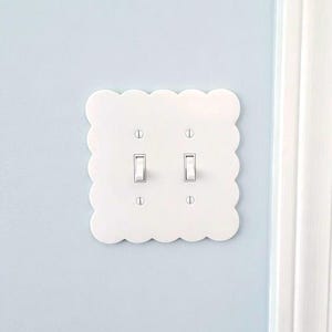 May include: White, scalloped-edge light switch cover with two toggle switches. The cover is mounted on a pale blue wall. A white door frame is visible on the right side of the image.
