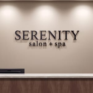 May include: A wall-mounted sign featuring the black, three-dimensional text SERENITY salon + spa centered on a smooth, light beige wall. Below the wall, a reception desk with a white stone countertop and a dark wood base is partially visible.
