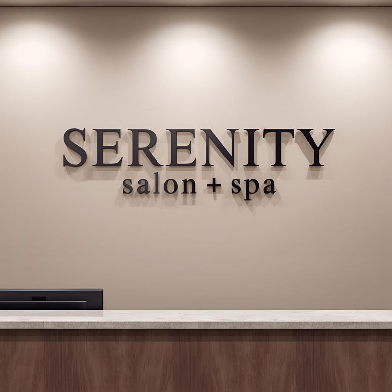 May include: A wall-mounted sign featuring the black, three-dimensional text SERENITY salon + spa centered on a smooth, light beige wall. Below the wall, a reception desk with a white stone countertop and a dark wood base is partially visible.