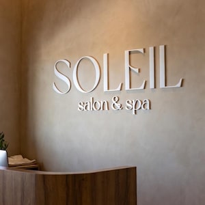 May include: A wall-mounted sign featuring the white, three-dimensional lettering "SOLEIL" in a large serif font, positioned above the smaller white text "salon & spa" on a textured, light beige wall surface above a curved wooden reception desk.