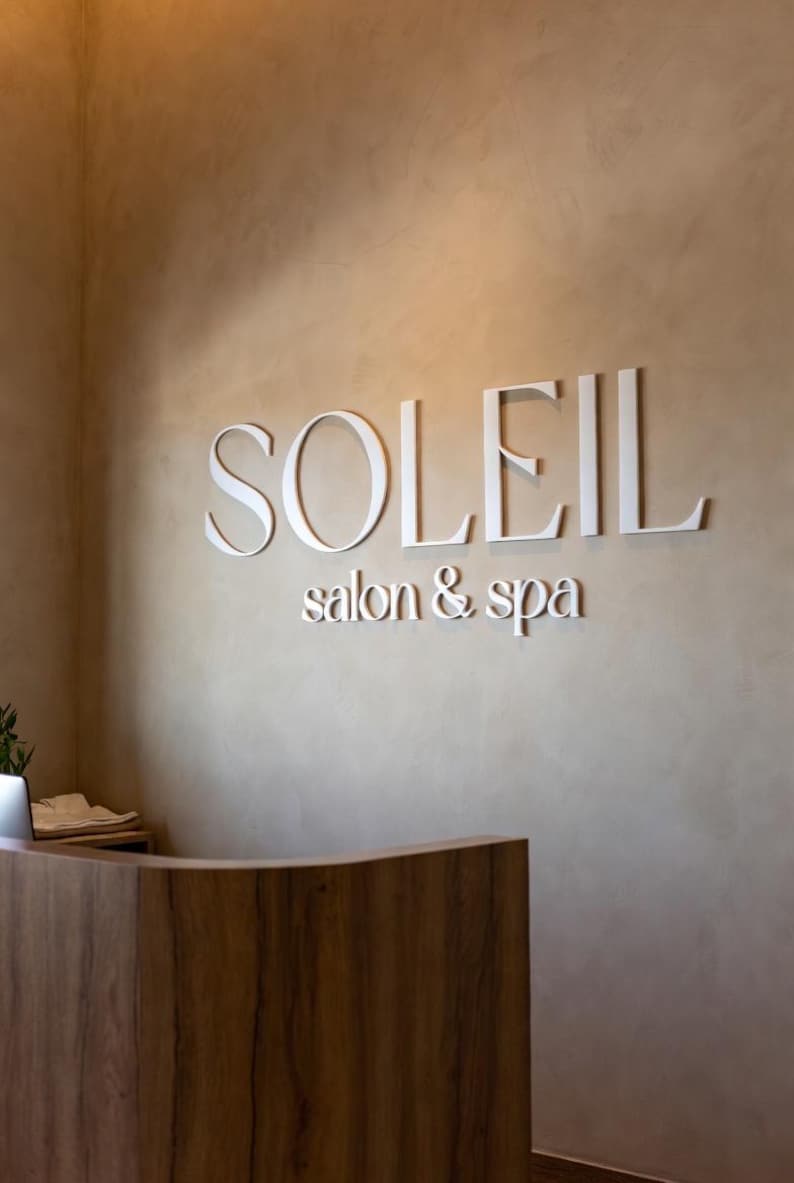 May include: A wall-mounted sign featuring the white, three-dimensional lettering "SOLEIL" in a large serif font, positioned above the smaller white text "salon & spa" on a textured, light beige wall surface above a curved wooden reception desk.