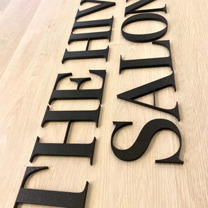 May include: A set of individual black, matte-finish, three-dimensional letters forming the words THE HIVE SALON. The letters are arranged in two vertical columns, featuring a classic serif typeface with clean, sharp edges and a textured surface.