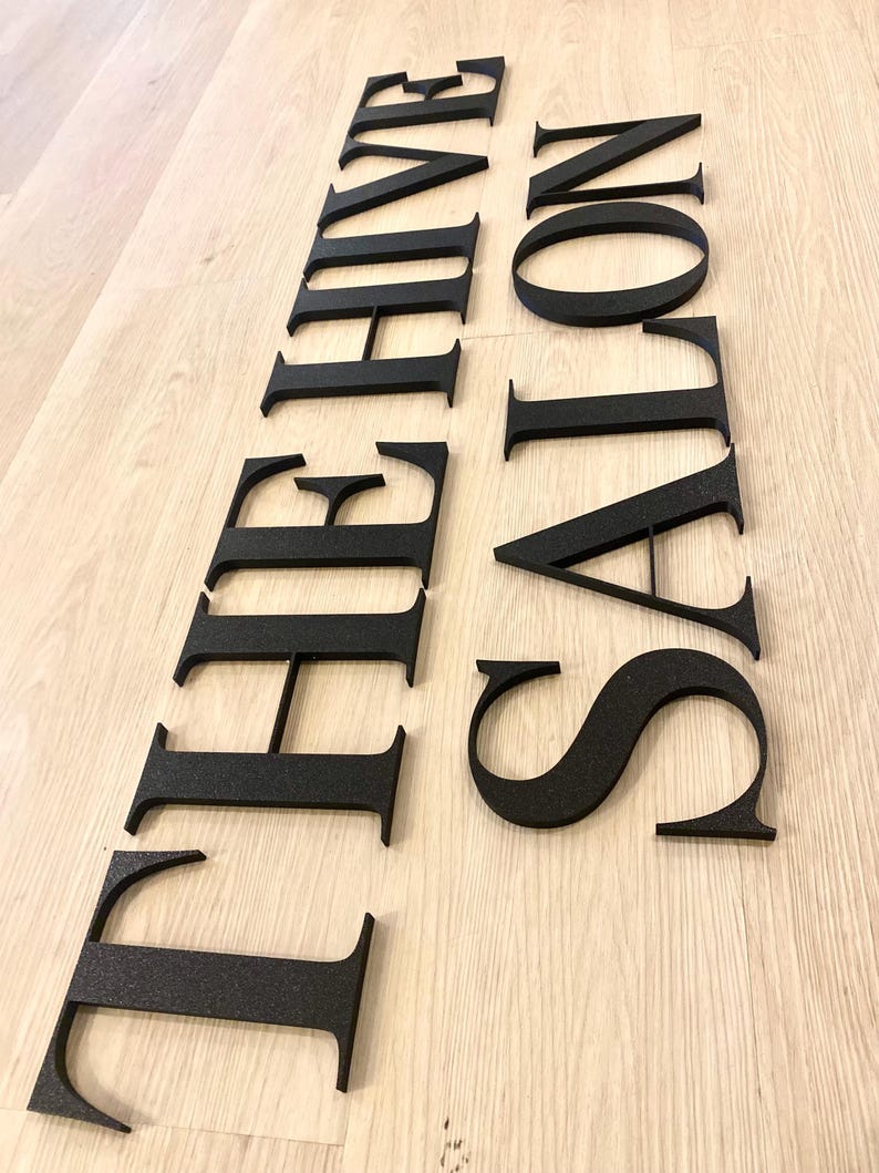 May include: A set of individual black, matte-finish, three-dimensional letters forming the words THE HIVE SALON. The letters are arranged in two vertical columns, featuring a classic serif typeface with clean, sharp edges and a textured surface.